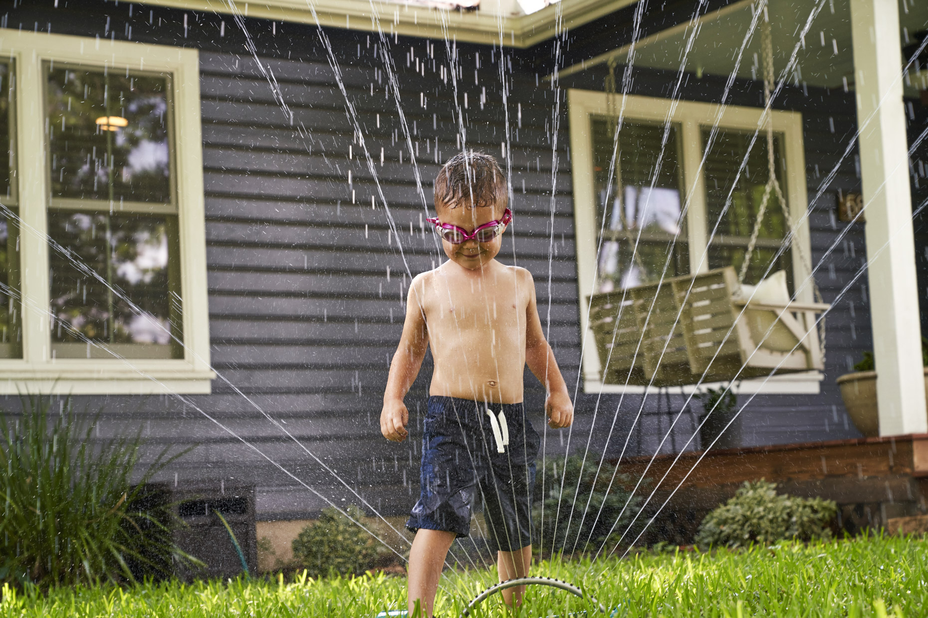 Boy in swim goggles playing in sprinkler in front yard