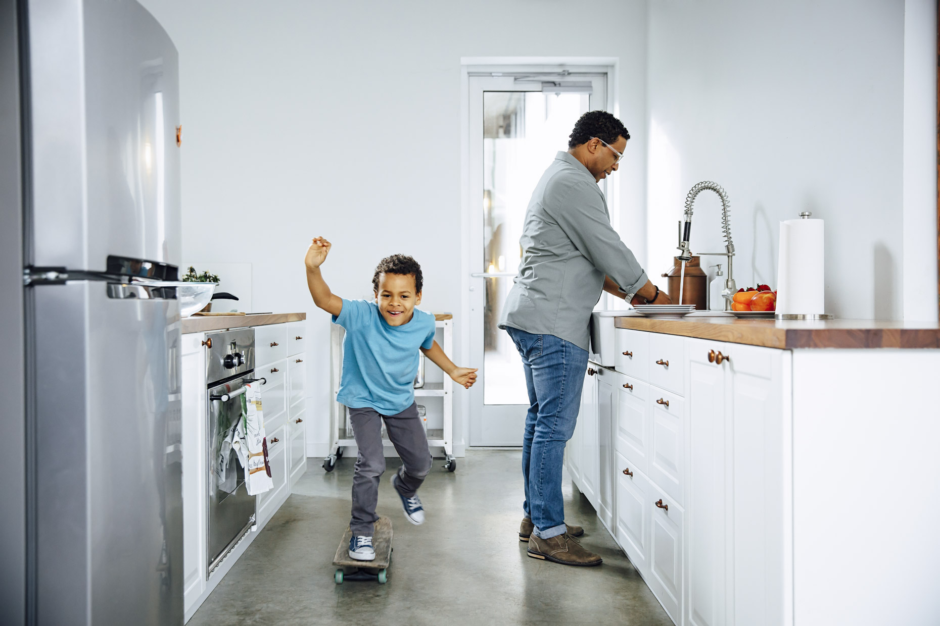 Boy skateboarding in kitchen while man does dishes