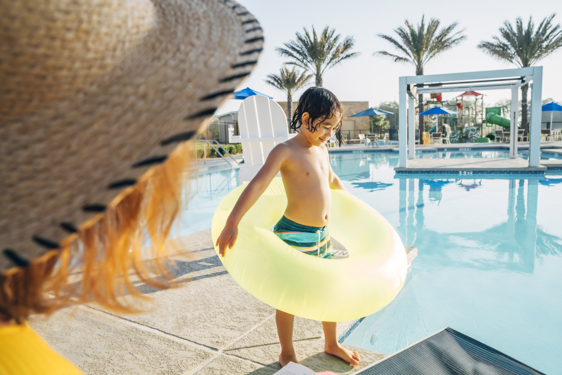Boy with yellow innertube walking to pool at resort while mom looks on