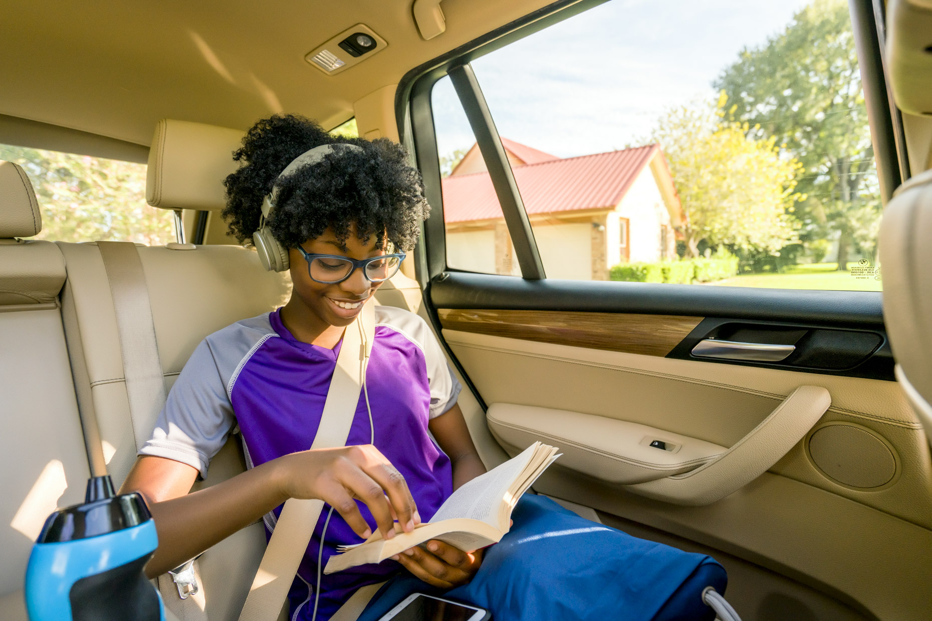 Girl in headphones and glasses reading book in back seat of car