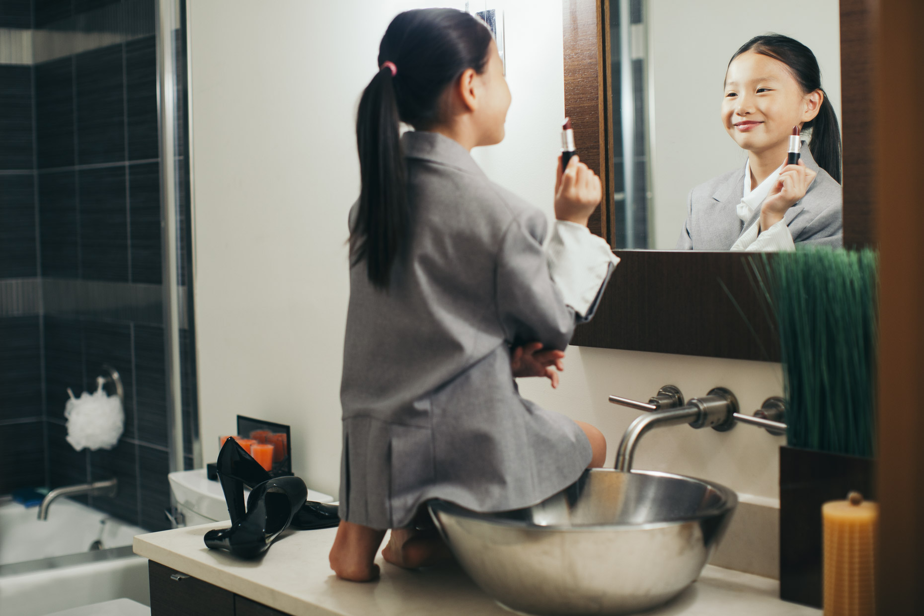 Asian girl in suit jacket on bathroom counter trying on lipstick in front of mirror
