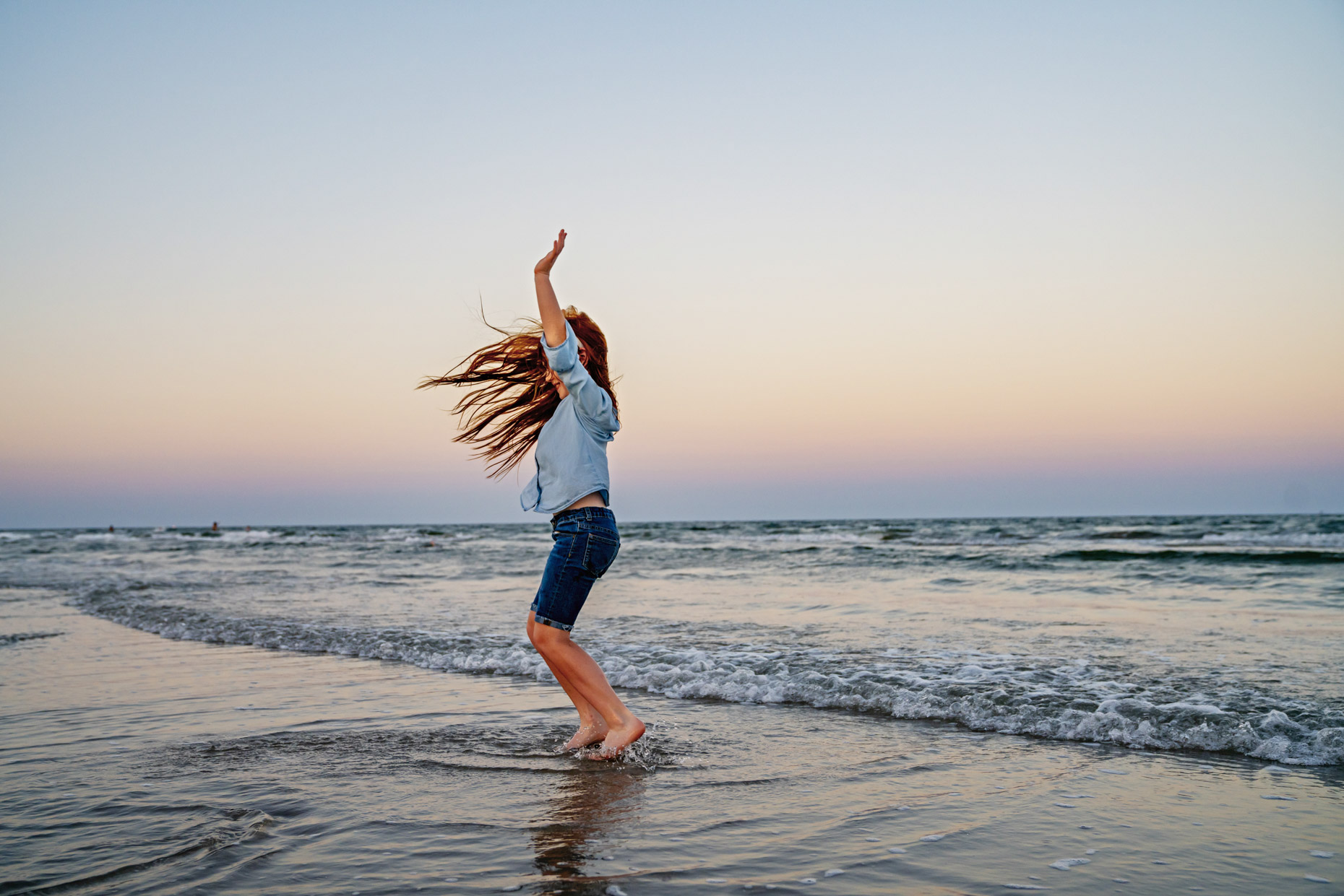 Girl with long red hair jumping in the waves on beach at sunset