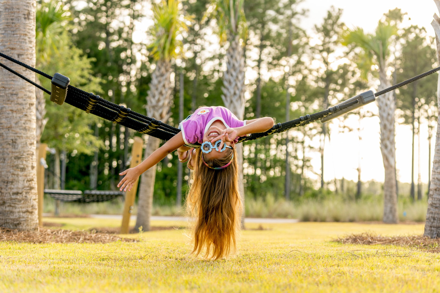 Girl-smiling-hanging-drom-hamock-upside-down-with-flower-glasses-and-peace-sign-042623_WILDLIGHT_SCENE7_9862