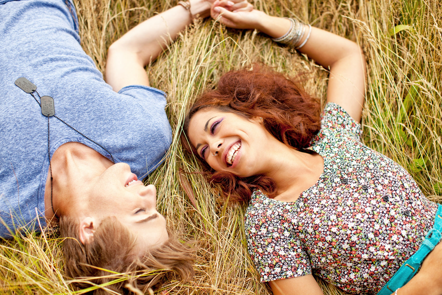 Happy teen couple laying down in grass outside holding hands looking at each other