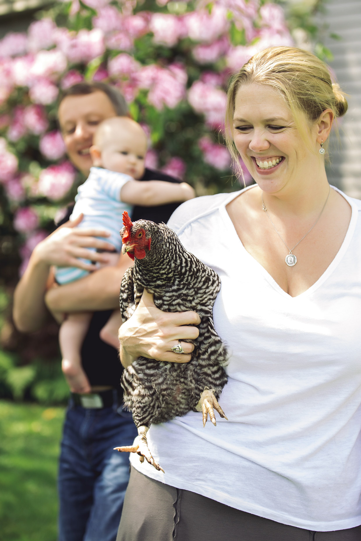 Happy woman holding chicken in garden with mand and baby walking behind