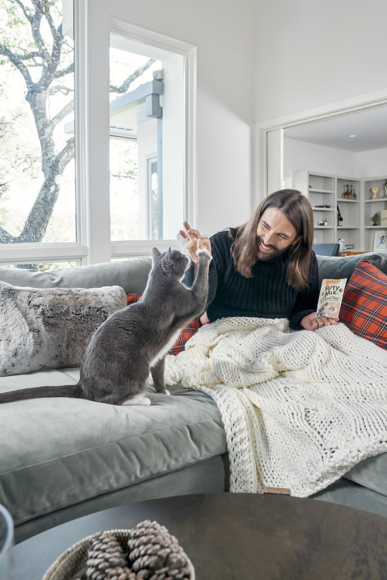 Jonathan Van Ness feeding cat Purina Friskies party mix treat on sofa