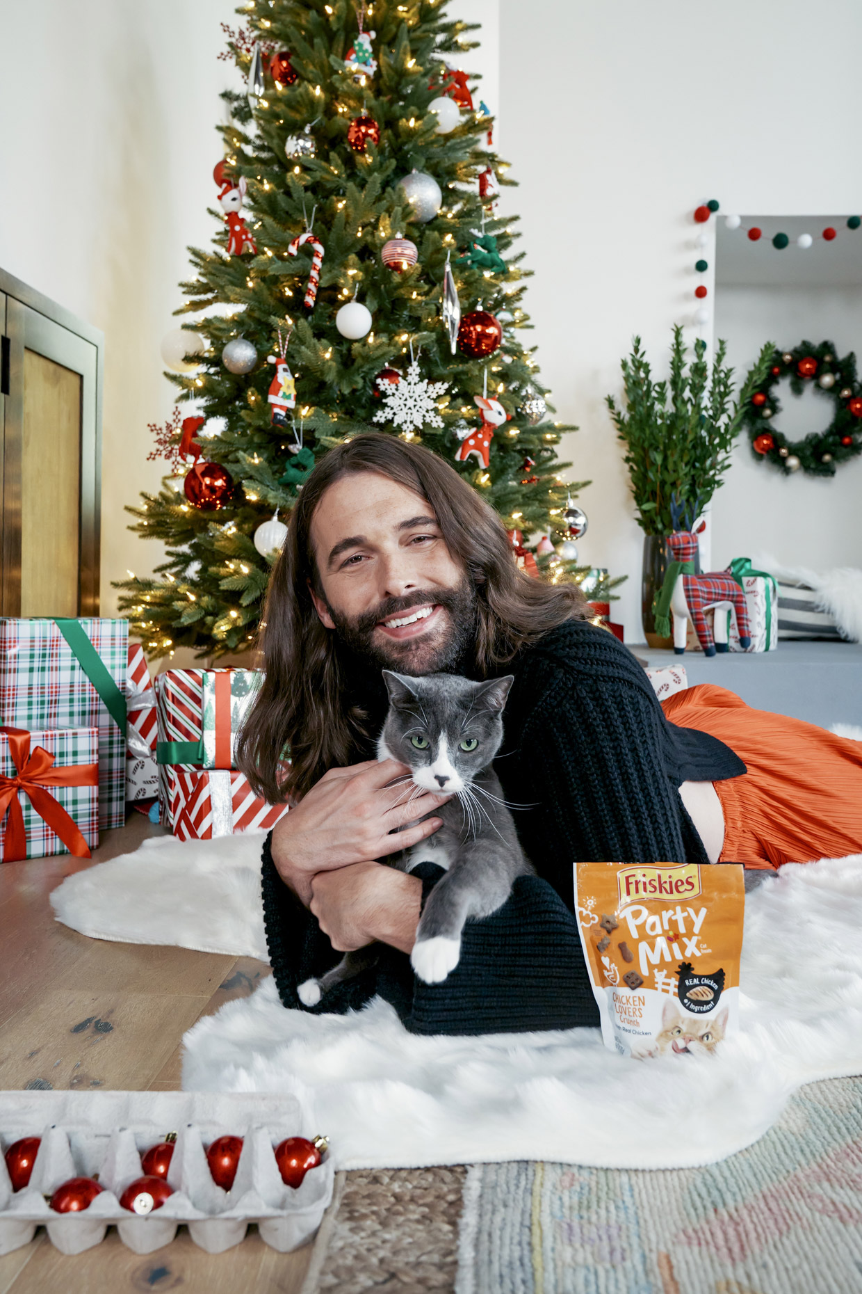 Jonathan Van Ness snuggling cat in front of Christmas tree with Friskies party mix