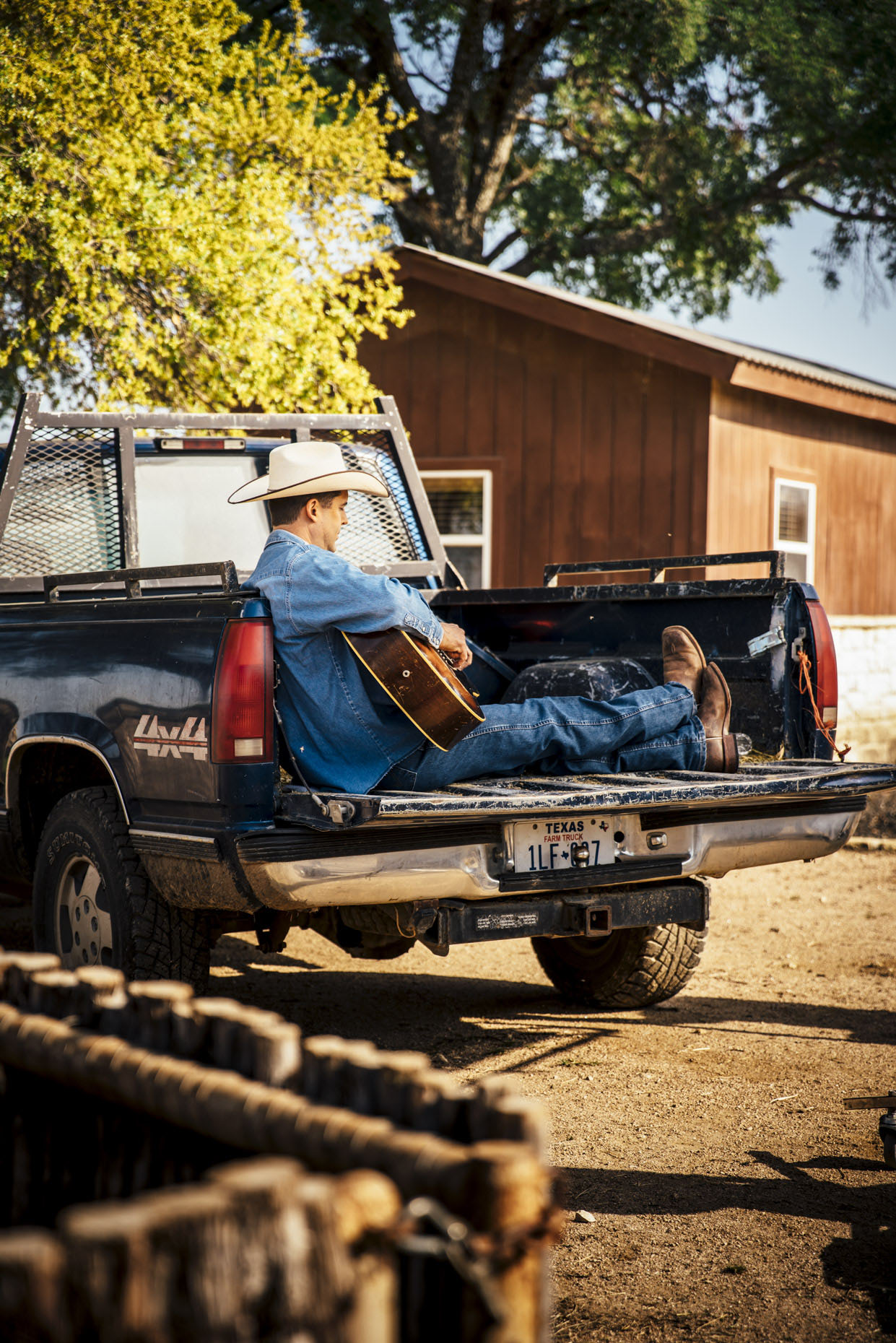 Man in cowboy hat sitting in back of pickup truck on horse ranch playing guitar