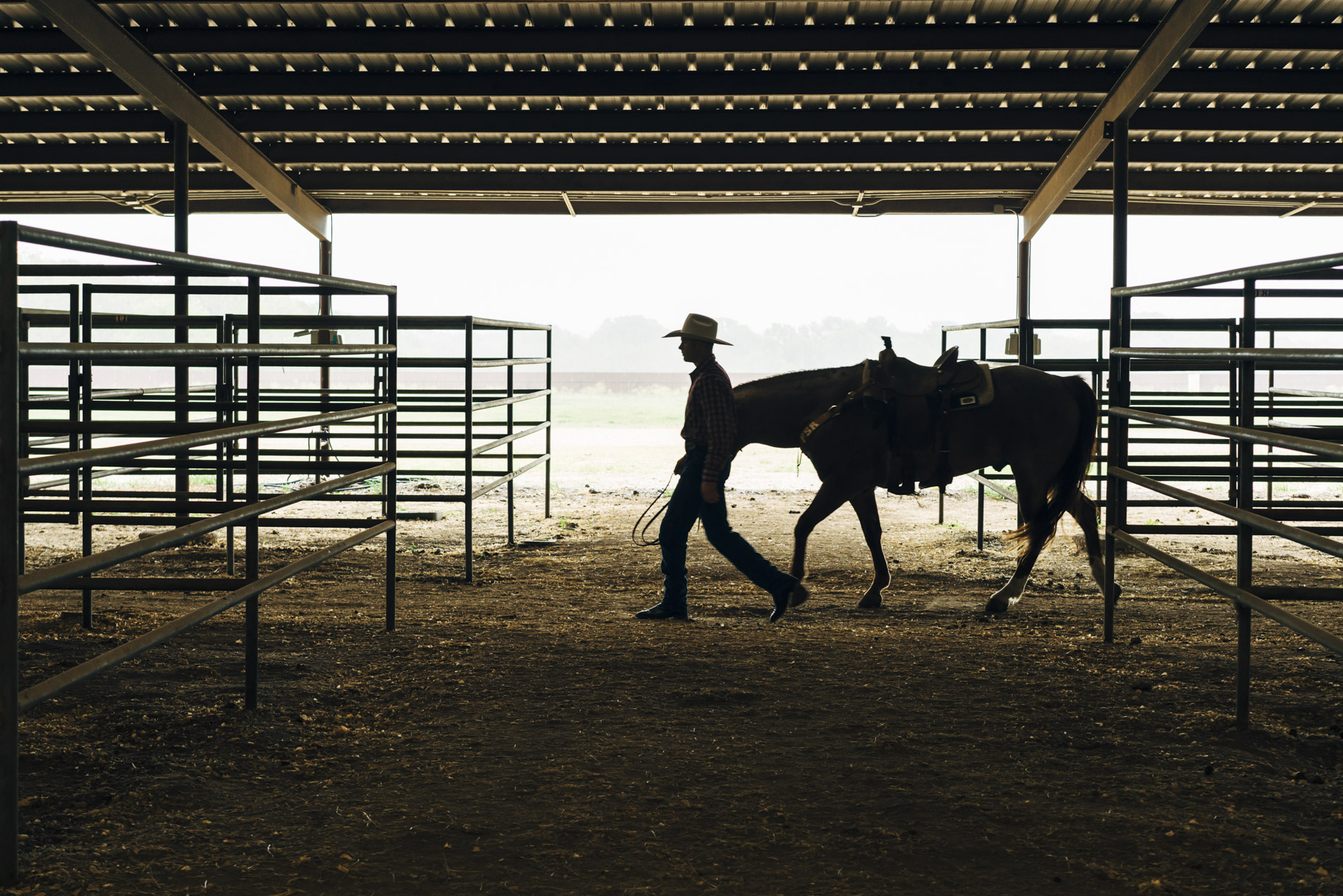 Silhouette of man in cowboy hat walking with horse through stable