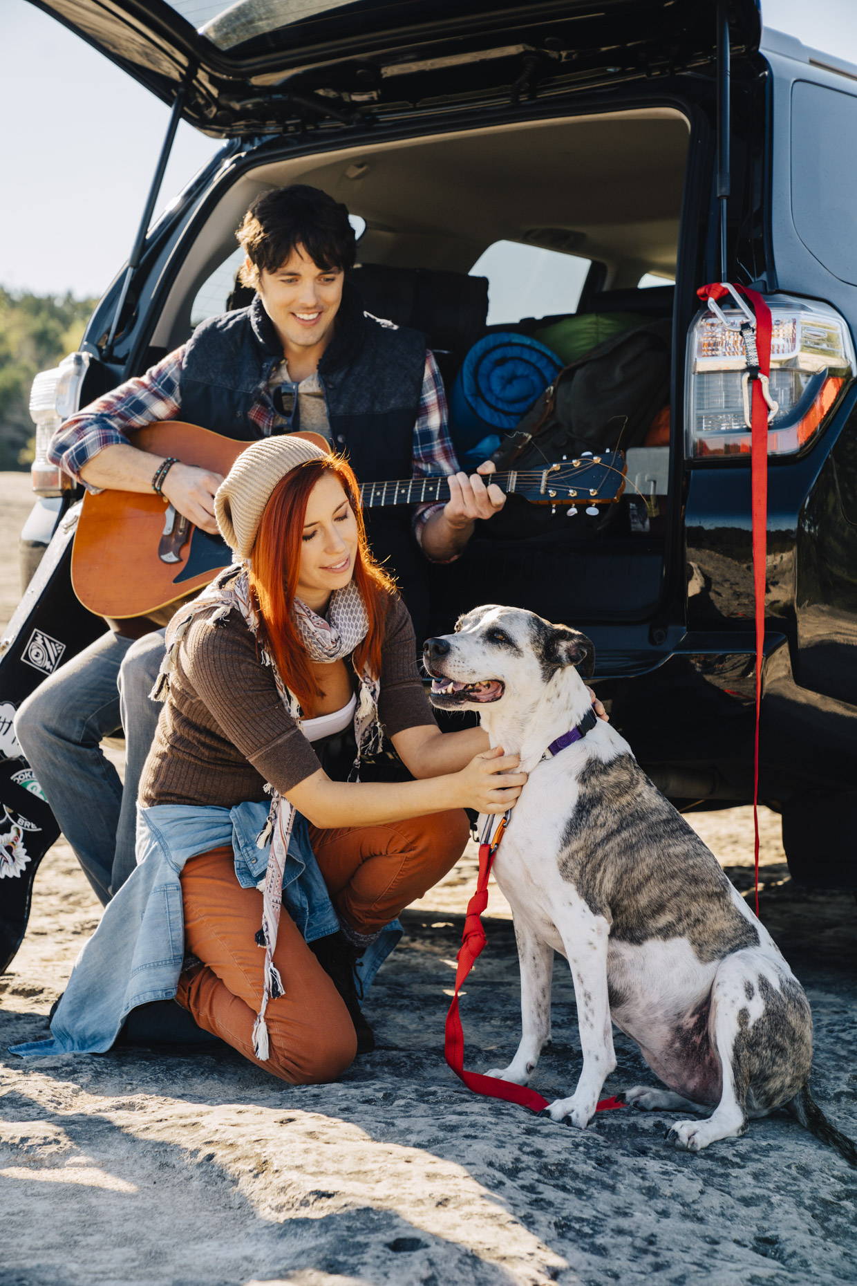 Man playing guitar in back of truck while woman pets dog