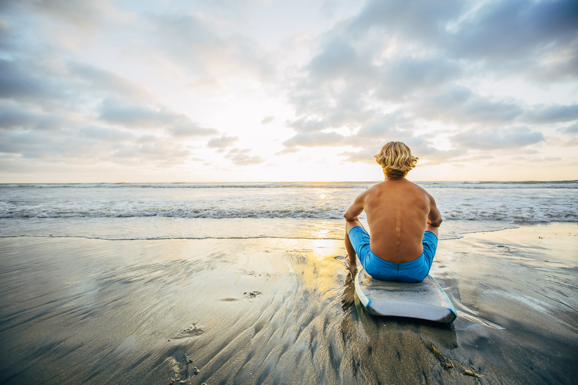 Man sitting on surfboard on beach looking out at ocean sunset