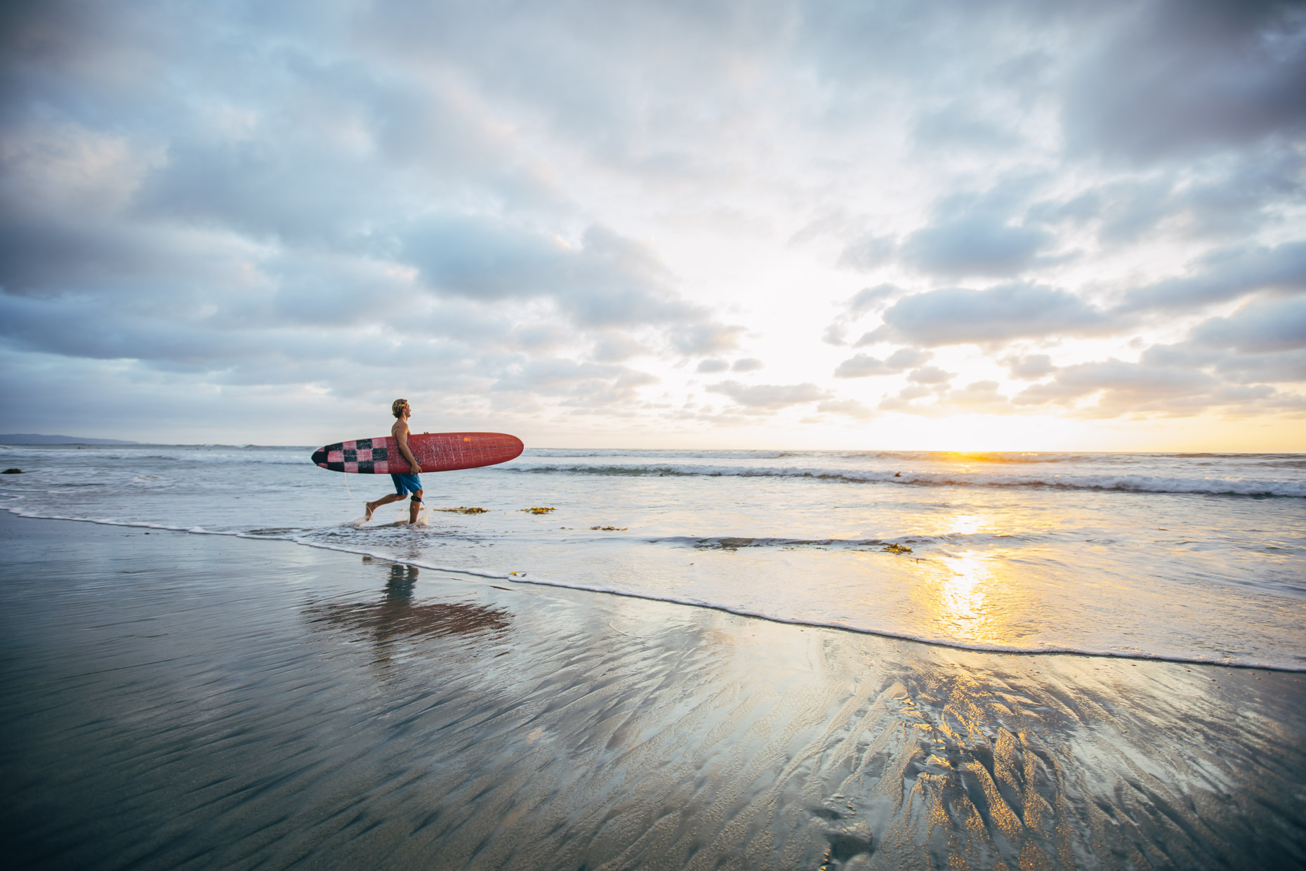 Man with surfboard running into ocean on beach at sunset