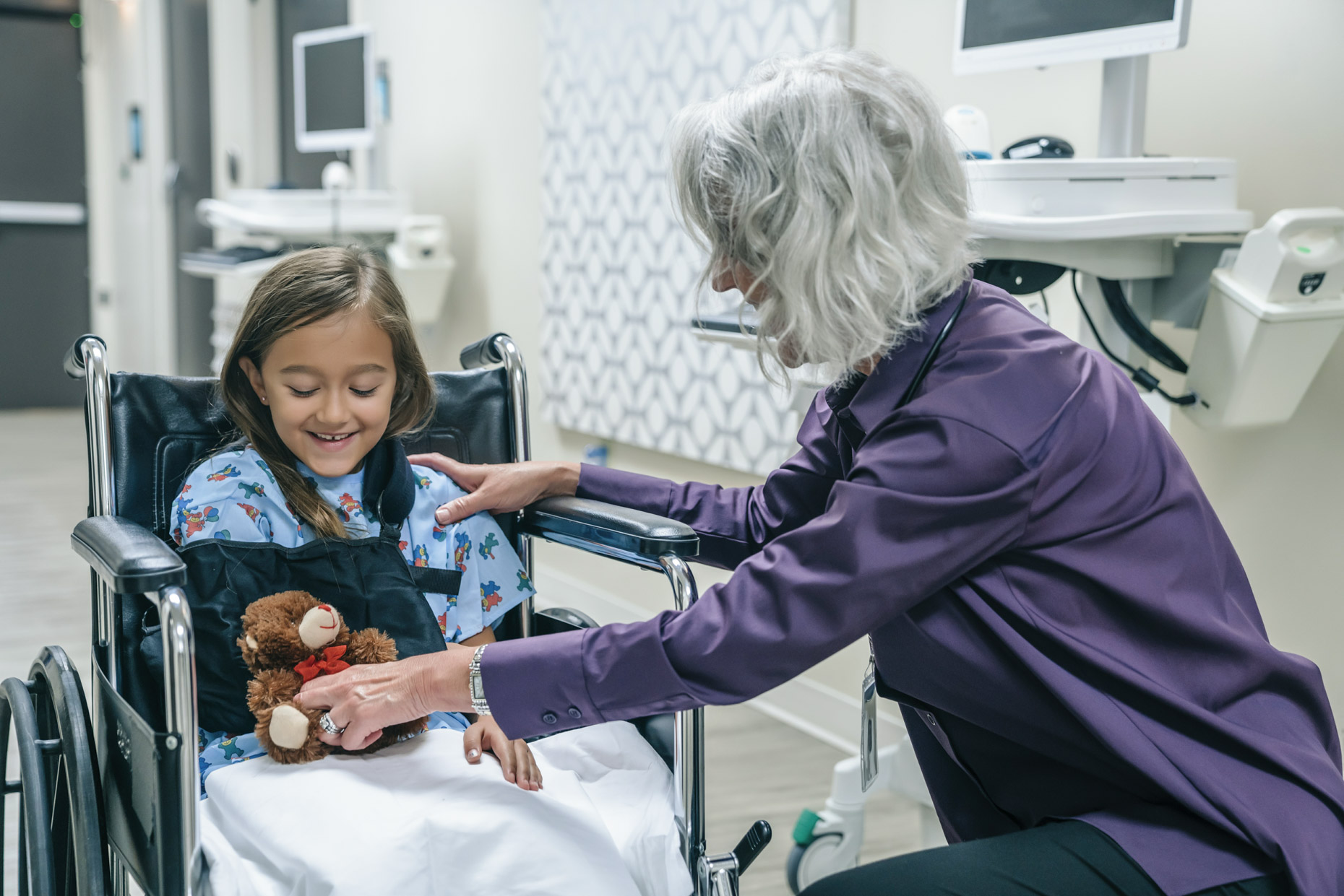 Mature female doctor giving teddy bear to girl in wheel chair with arm in sling