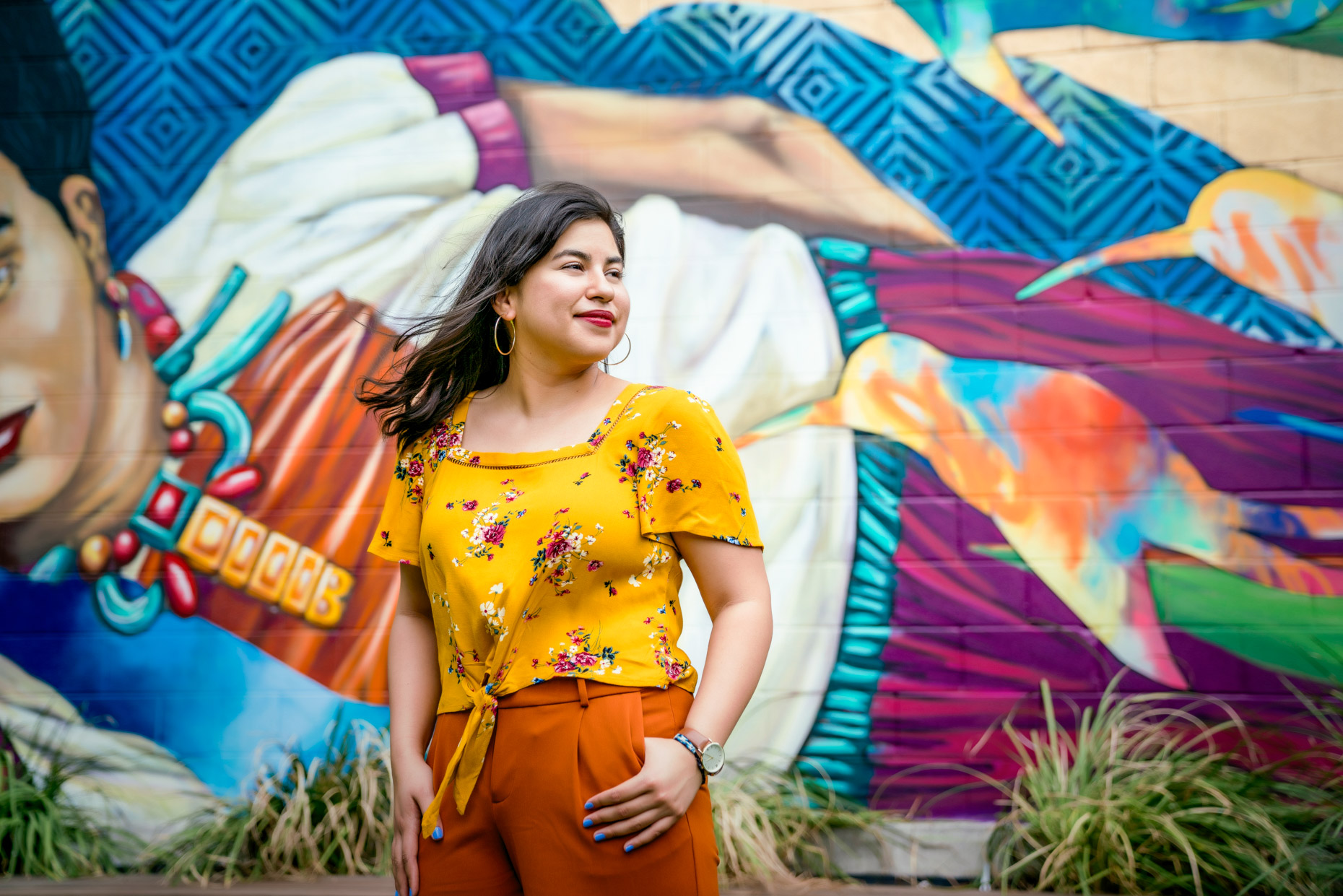 Portrait profile of latina woman in floral top in front of brightly colored Frida Kahlo mural