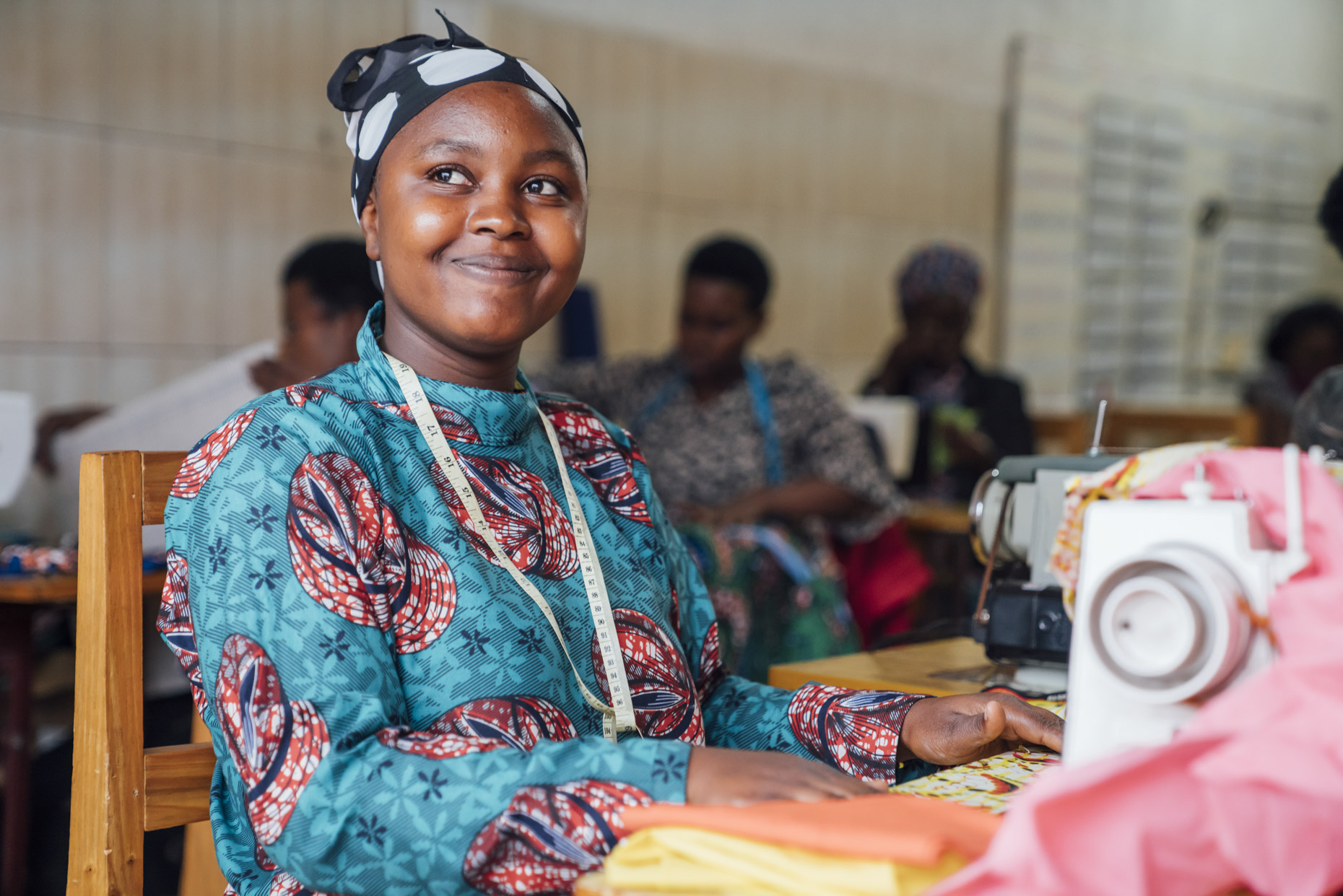 Seamstress in bright clothes sitting at sewing maching looking up and smiling