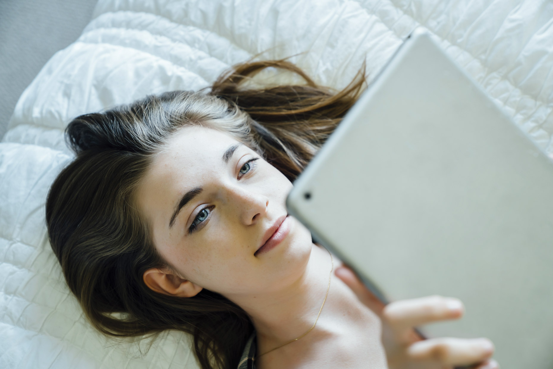 Teen girl with blue eyes laying on bed looking at ipad tablet computer
