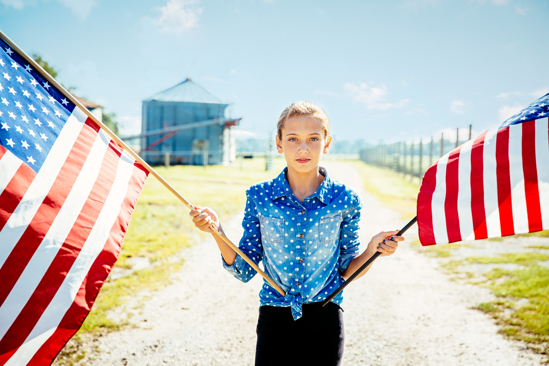 Teen girl holding american flags in front of grain silo on ranch