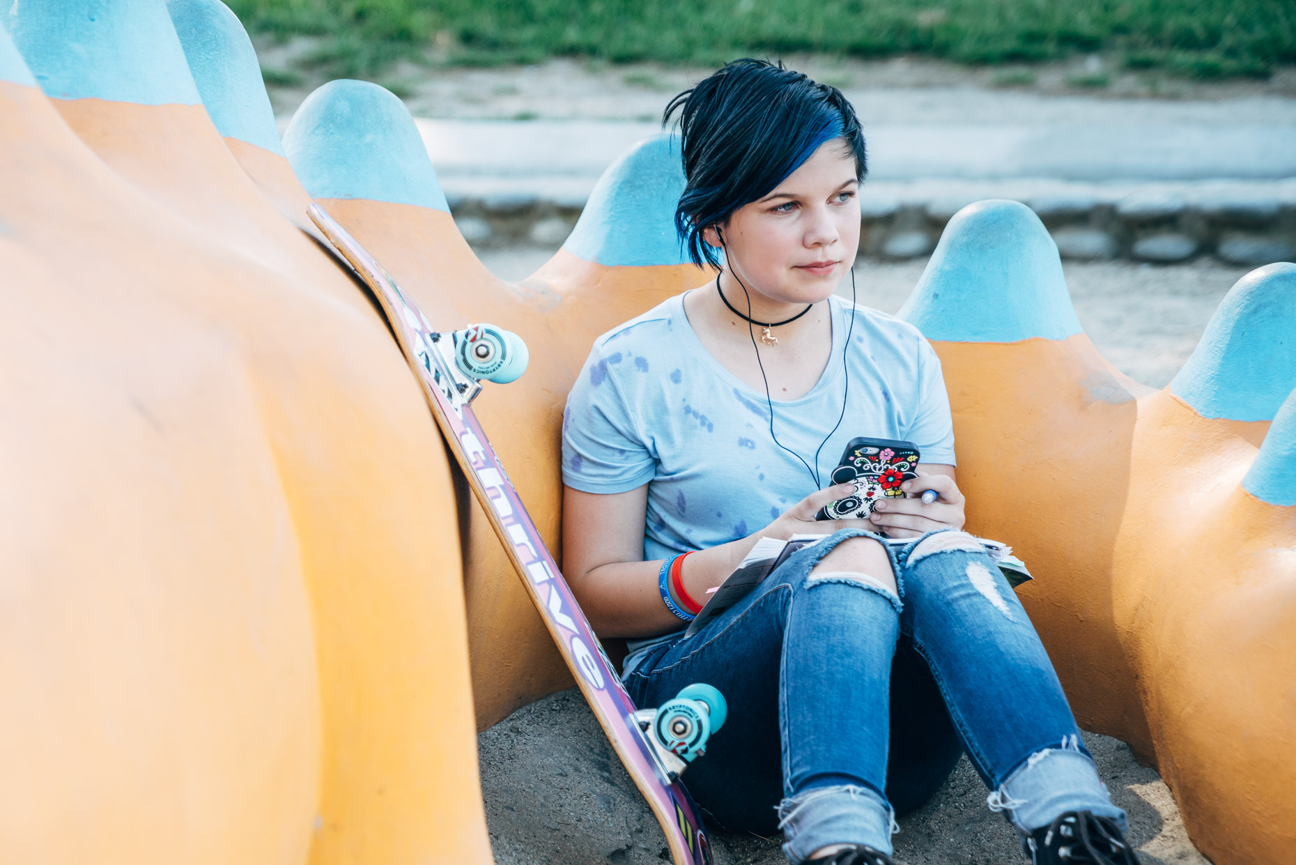 Transgender teen with skate board, cell phone, and headphones sitting in park journaling