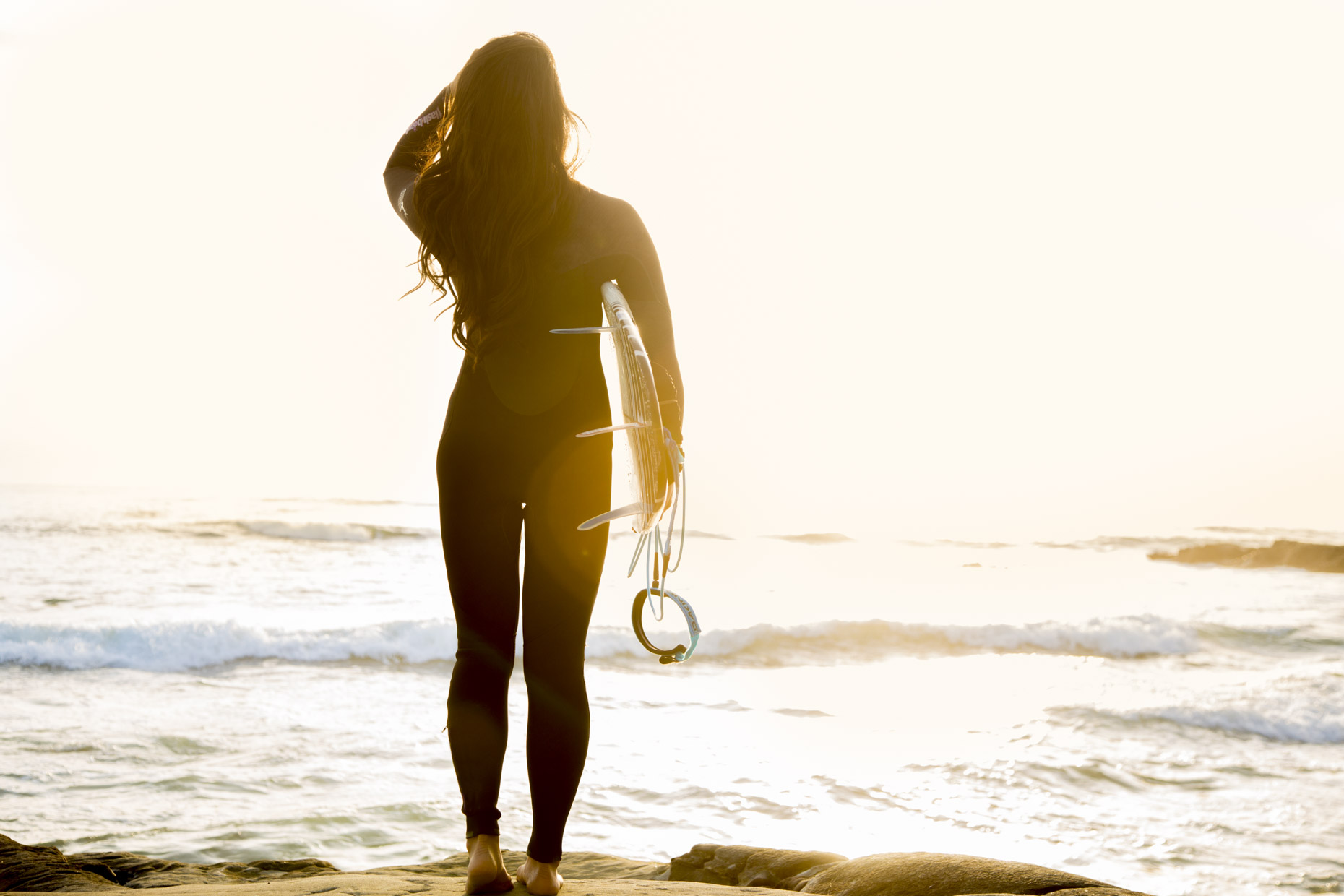 Woman in wetsuit holding surfboard looking at ocean at sunset