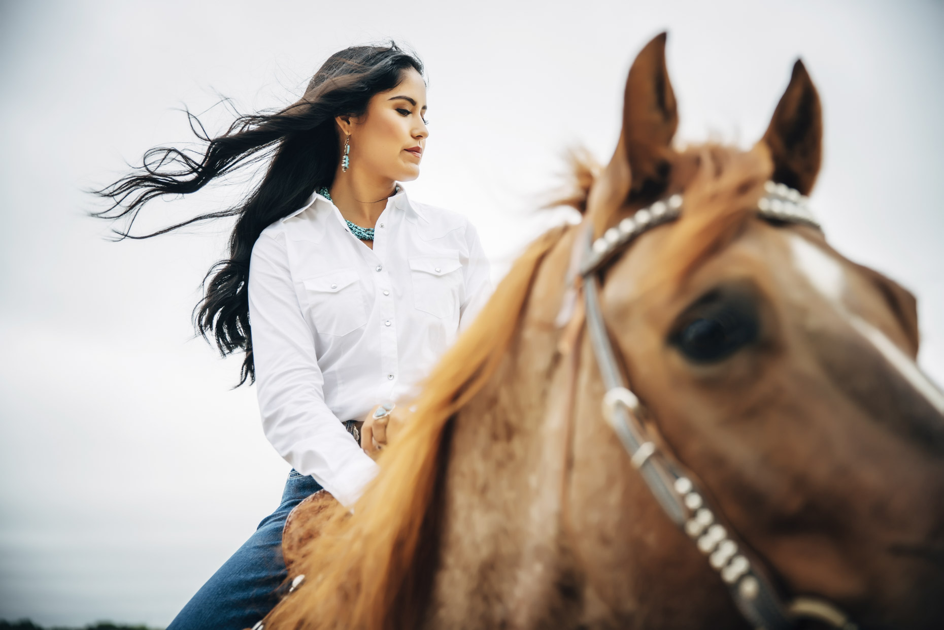 Woman iwith long brown hair in white Wrangler shirt riding horse