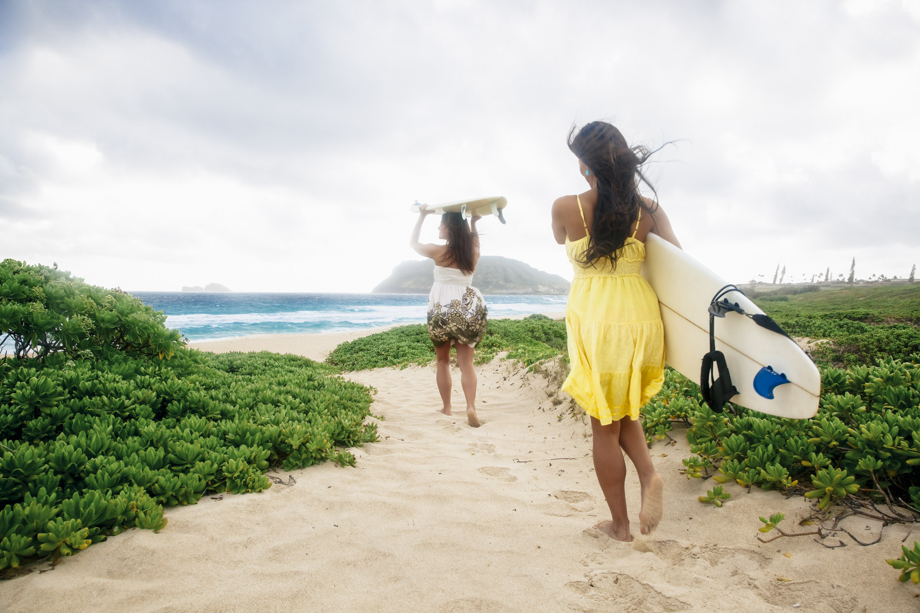 Women in dresses walking toward ocean with surfboards