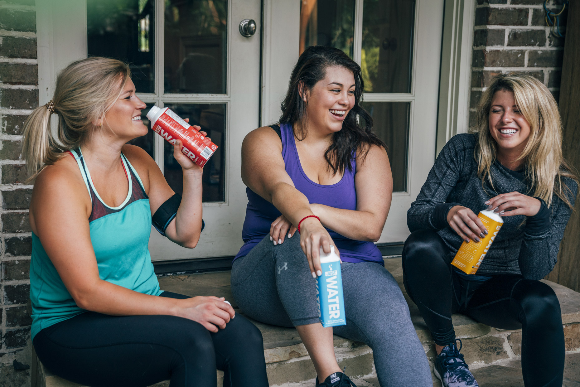 Women sitting on steps outside fitness studio drinking boxed water and laughing