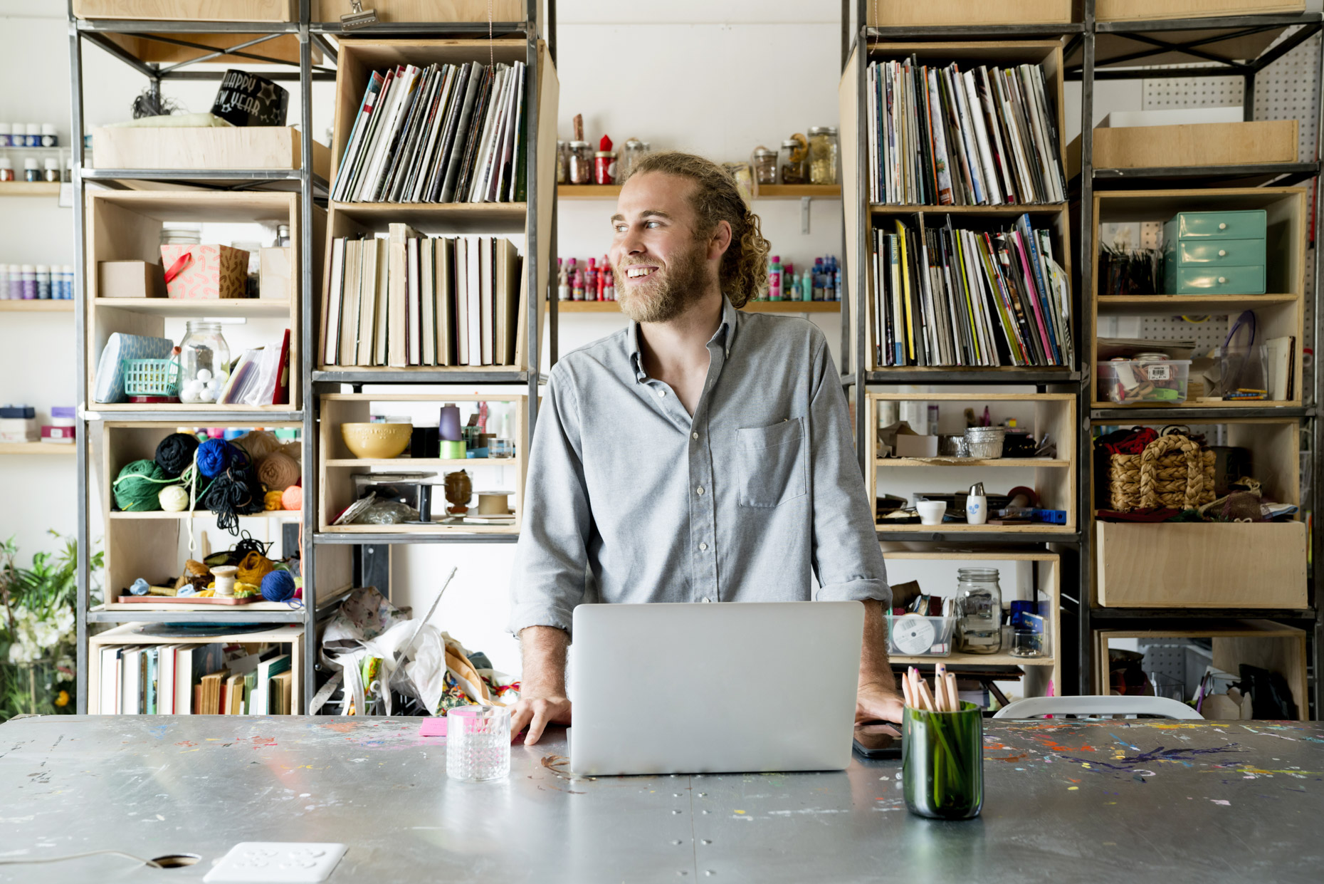 Profile of happy bearded man with long hair in bun standing in front of craft space with laptop