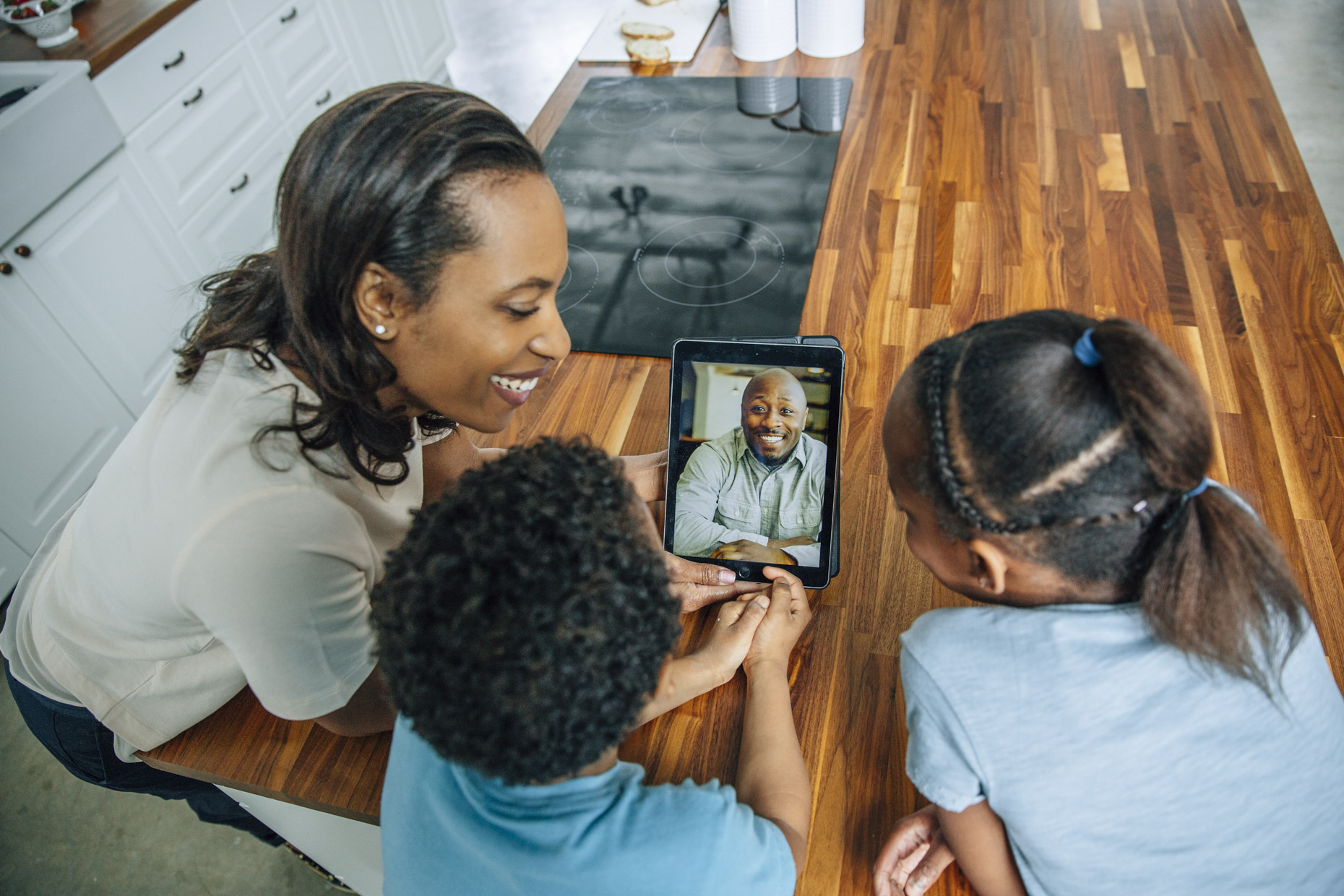Woman and kids video chatting with dad on iPad tabled computer in kitchen